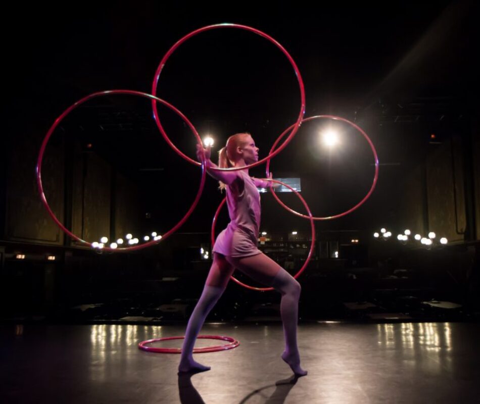 Annika Hakala hulahoop artist performing on theatre stage in Vienna with four magenta hulahoops. Dark background and pink tones. Annika is in elegant pose and the hulahoops are circling around her both arms.