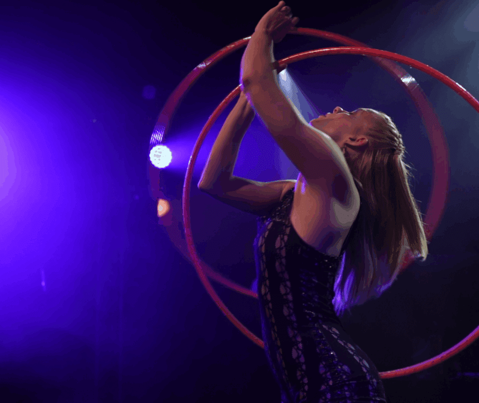 Hulahoop artist Annika Hakala pictured from side manipulating two pink hula hoops with her hands around her upper body. Shot in a performance on theater stage. Elegant mesmerising atmosphere in dark lilac stage lights.