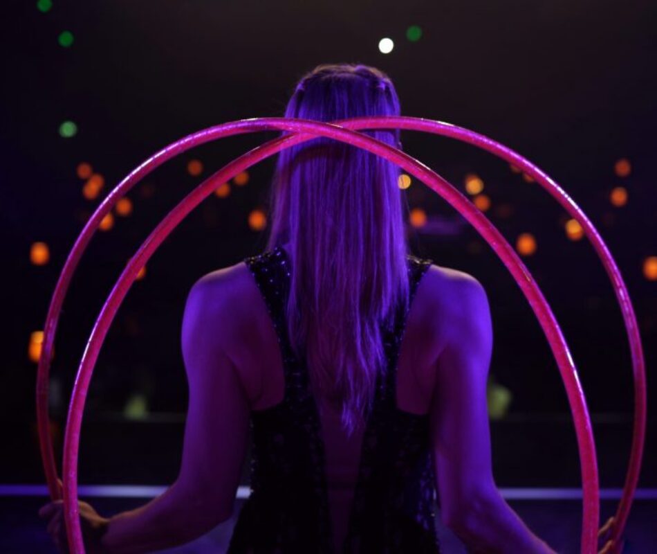 Hulahoop artist Annika Hakala pictured from behind manipulating two pink hula hoops with her hands behind her back. Stage shot on a theatre stage from behind audience room on background. Elegant mesmerising atmosphere in dark lilac stage lights and dark background.