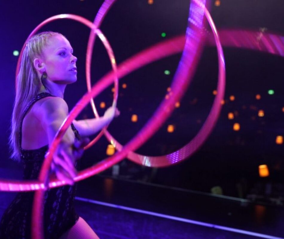 Hulahoop artist Annika Hakala pictured from side manipulating four pink hulahoops with her hands. Stageshot on a theatre stage audience room as a background. Elegant mesmerising atmosphere in dark lilac stage lights and dark background.