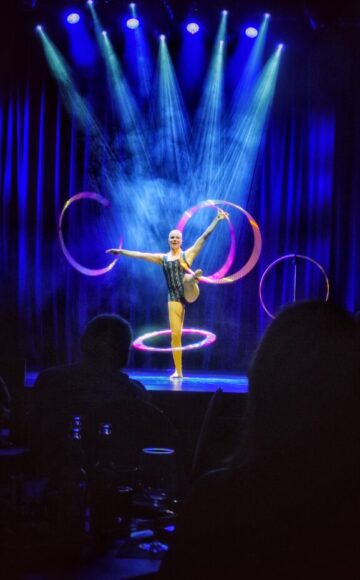 Circus artist Annika Hakala standing on stage on one feet and turning hula hoops on her other foot and on her both arms. Blue background, pink hula hoops and audience at front.